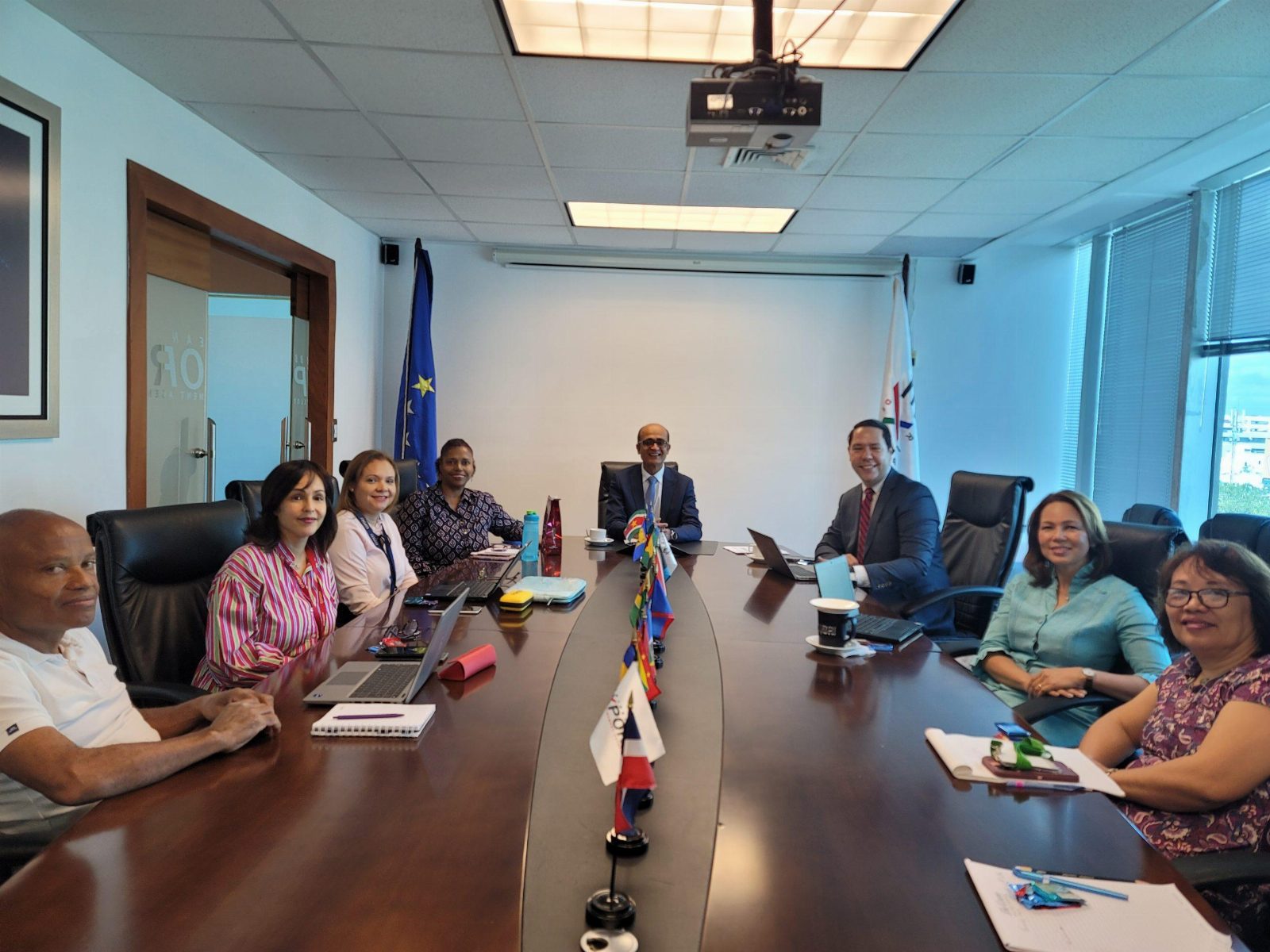 Eight colleagues seated around a boardroom table in a working meeting, with laptops, notepads and national flags in front of them.